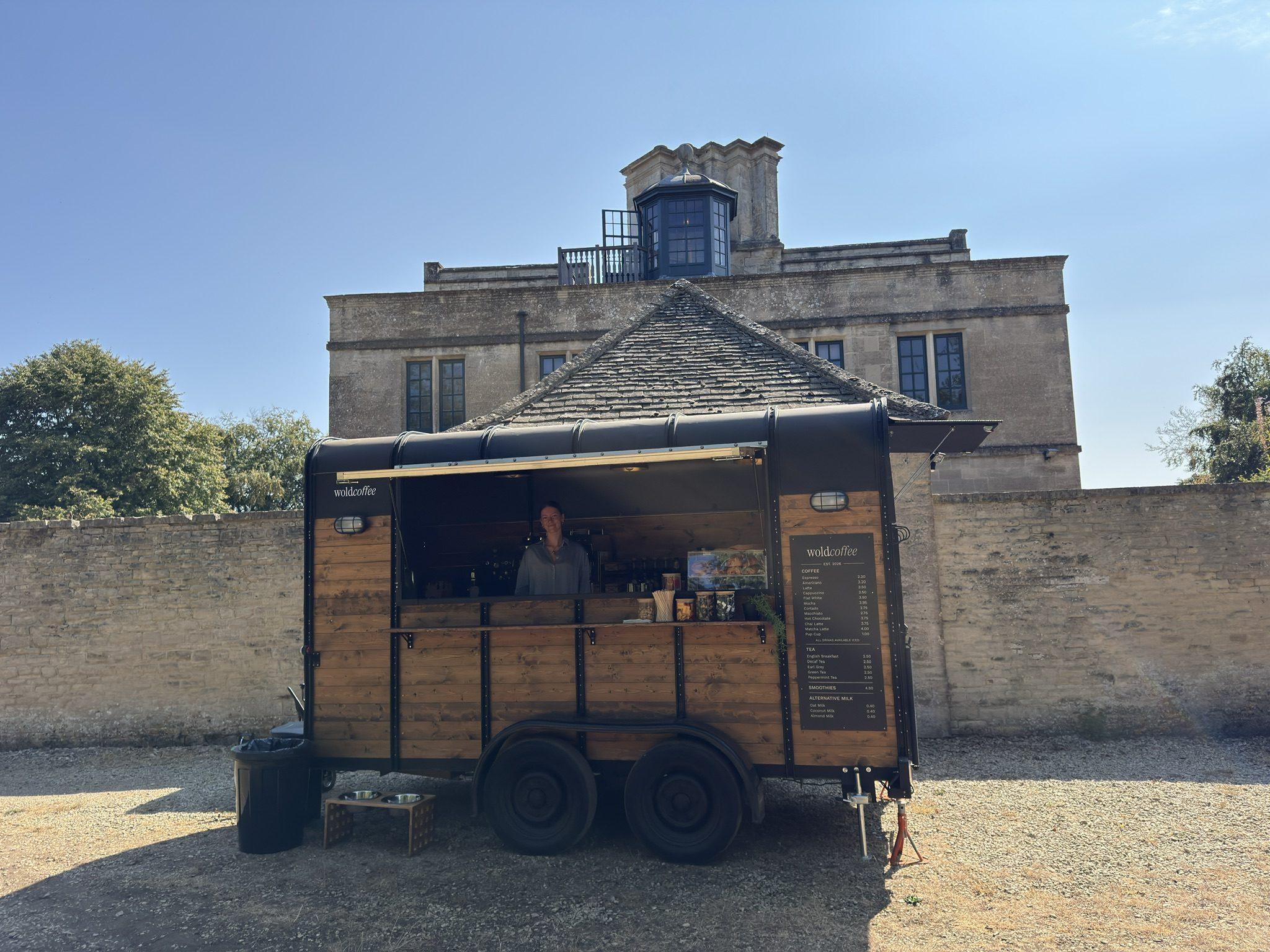 The WoldCoffee events trailer, a converted wooden horsebox with a black roof, pitched on gravel with its serving hatch propped open and a barista at the counter. Behind it stands a Cotswold stone house with a cupola on the roof.